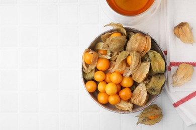 Ripe physalis fruits with calyxes in bowl on white tiled table, top view. Space for text