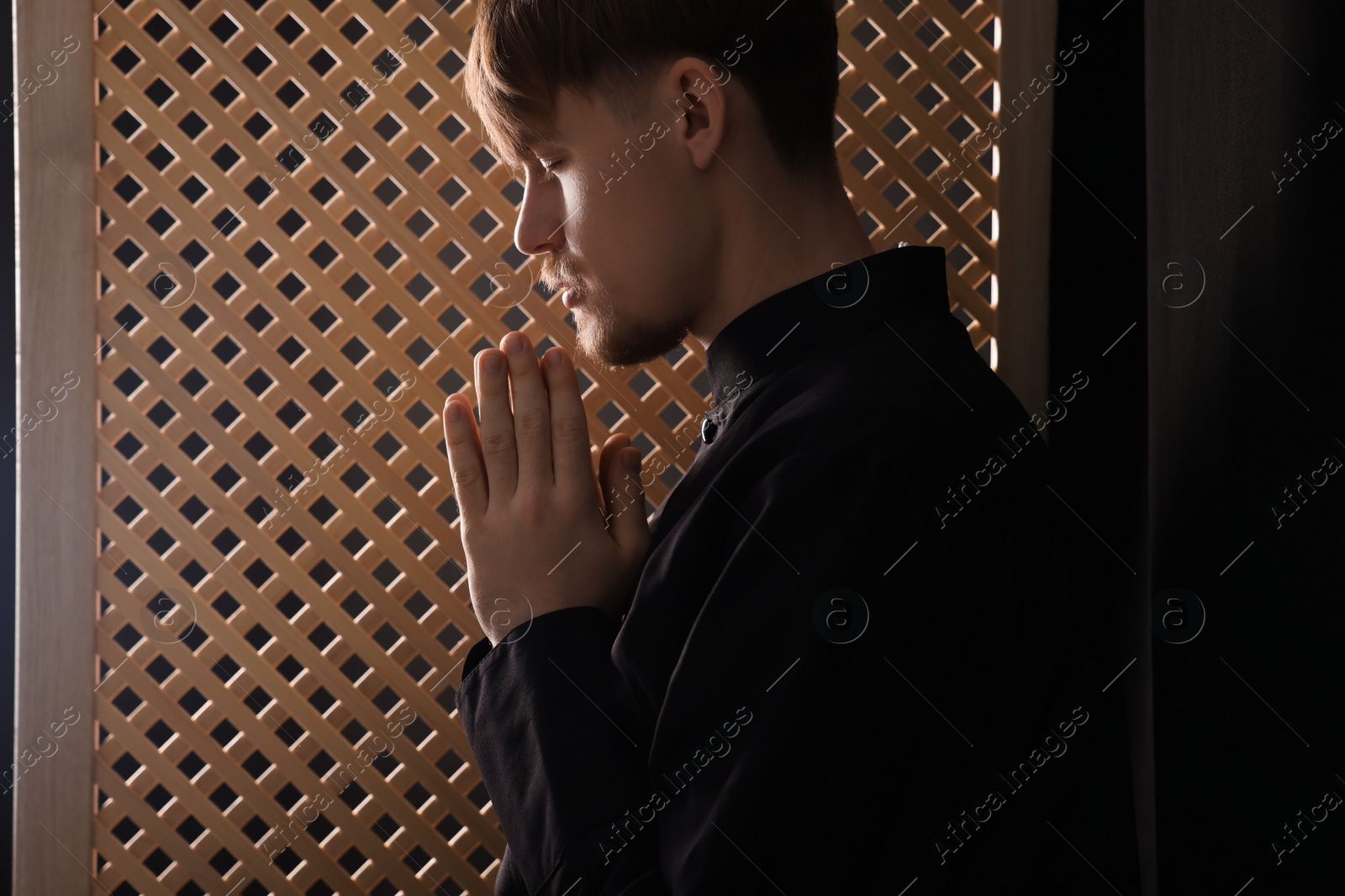 Photo of Catholic priest praying near wooden window in confessional booth