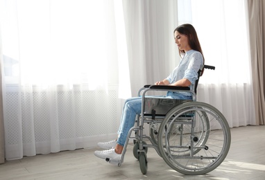 Young woman sitting in wheelchair near window indoors. Space for text