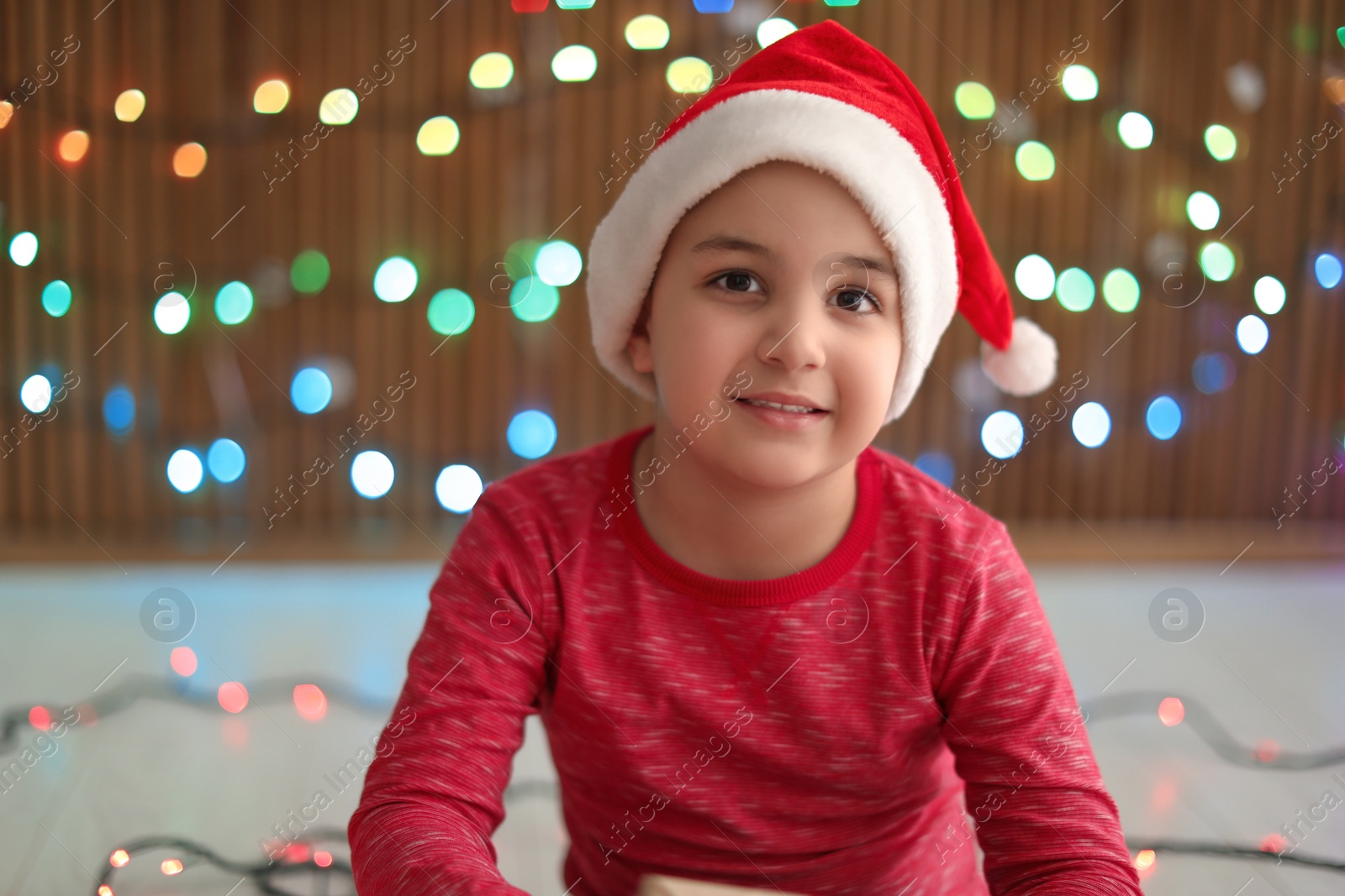 Photo of Cute little child in Santa hat on blurred lights background. Christmas celebration