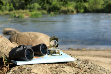 Binoculars, compass and map on stone near river, space for text. Camping equipment