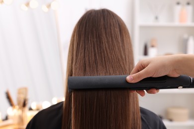 Photo of Hairdresser straightening woman's hair with flat iron in salon, closeup