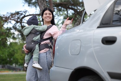 Photo of Mother holding her child in sling (baby carrier) while putting shopping bags into car trunk outdoors