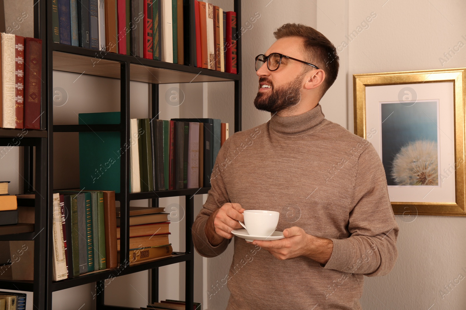 Photo of Young man with cup of coffee near shelves in home library