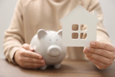 Photo of Man holding house model and piggy bank at wooden table, closeup. Saving money concept