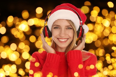 Image of Happy woman in Santa hat listening to Christmas music via headphones on dark background with blurred lights