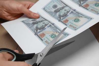 Photo of Counterfeiter cutting dollar banknotes with scissors at wooden table, closeup. Fake money concept