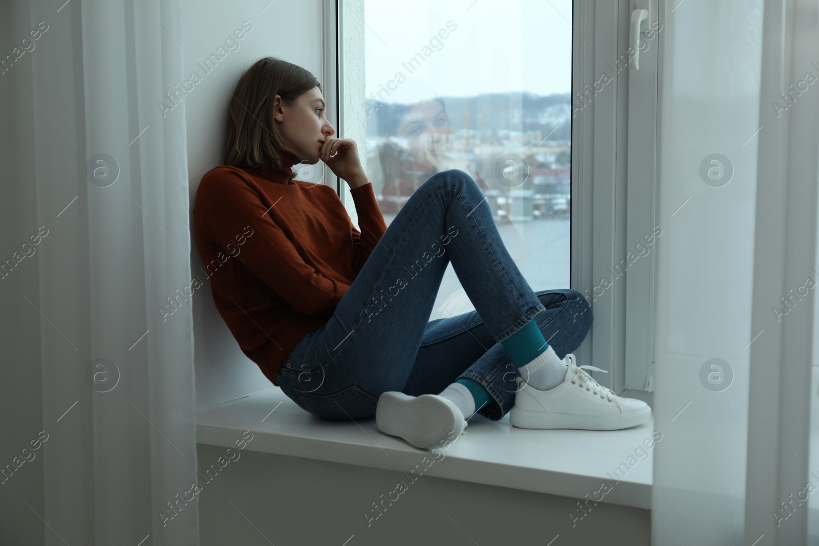 Photo of Sad young woman sitting on windowsill near window at home