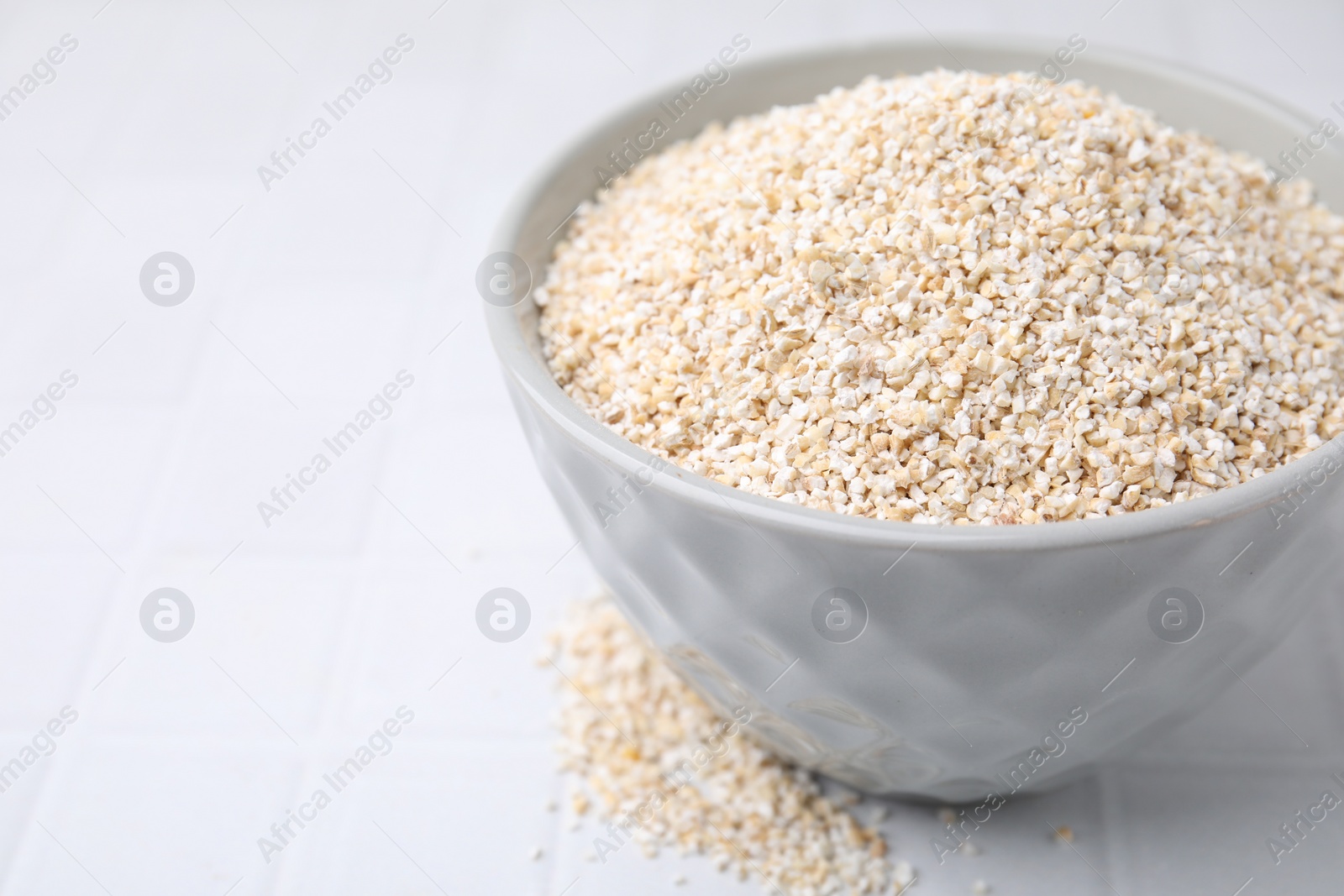 Photo of Dry barley groats in bowl on white tiled table, closeup. Space for text