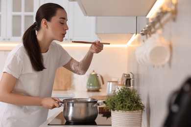 Beautiful woman with wooden spoon tasting soup in kitchen