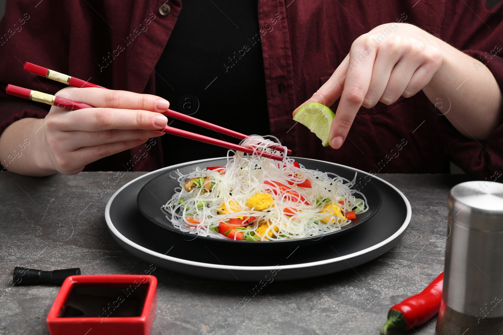 Photo of Stir-fry. Woman squeezing lime into tasty rice noodles with meat and vegetables at grey textured table, closeup