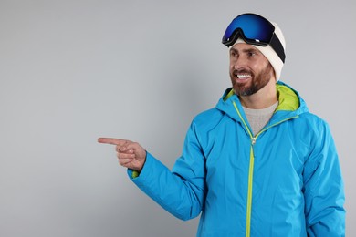 Winter sports. Happy man in ski suit and goggles pointing at something on gray background, space for text