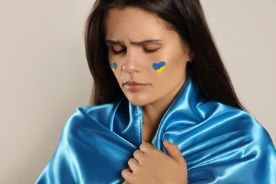 Sad young woman with face paint and Ukrainian flag on beige background, closeup