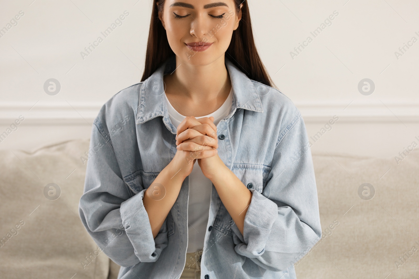 Photo of Religious young woman with clasped hands praying indoors, closeup