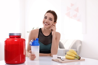 Young woman holding bottle of protein shake at table with ingredients in room. Space for text