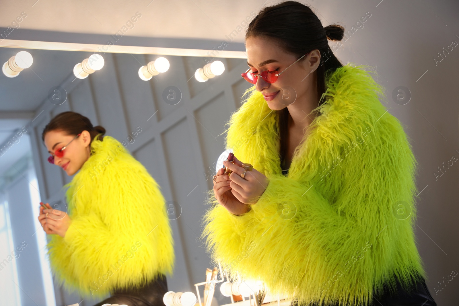 Photo of Young woman applying make up near illuminated mirror indoors