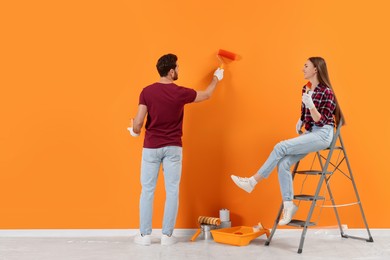Photo of Man painting orange wall and woman sitting on folding ladder indoors. Interior design
