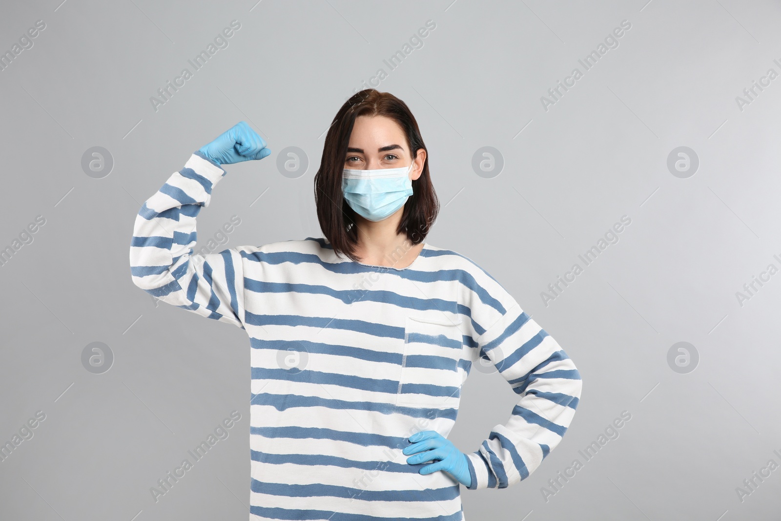 Photo of Woman with protective mask and gloves showing muscles on light grey background. Strong immunity concept