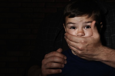 Adult man covering scared little boy's mouth on dark background, space for text. Child in danger