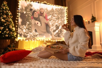 Image of Woman eating tasty pizza and watching romantic movie via video projector in room. Cozy winter holidays atmosphere