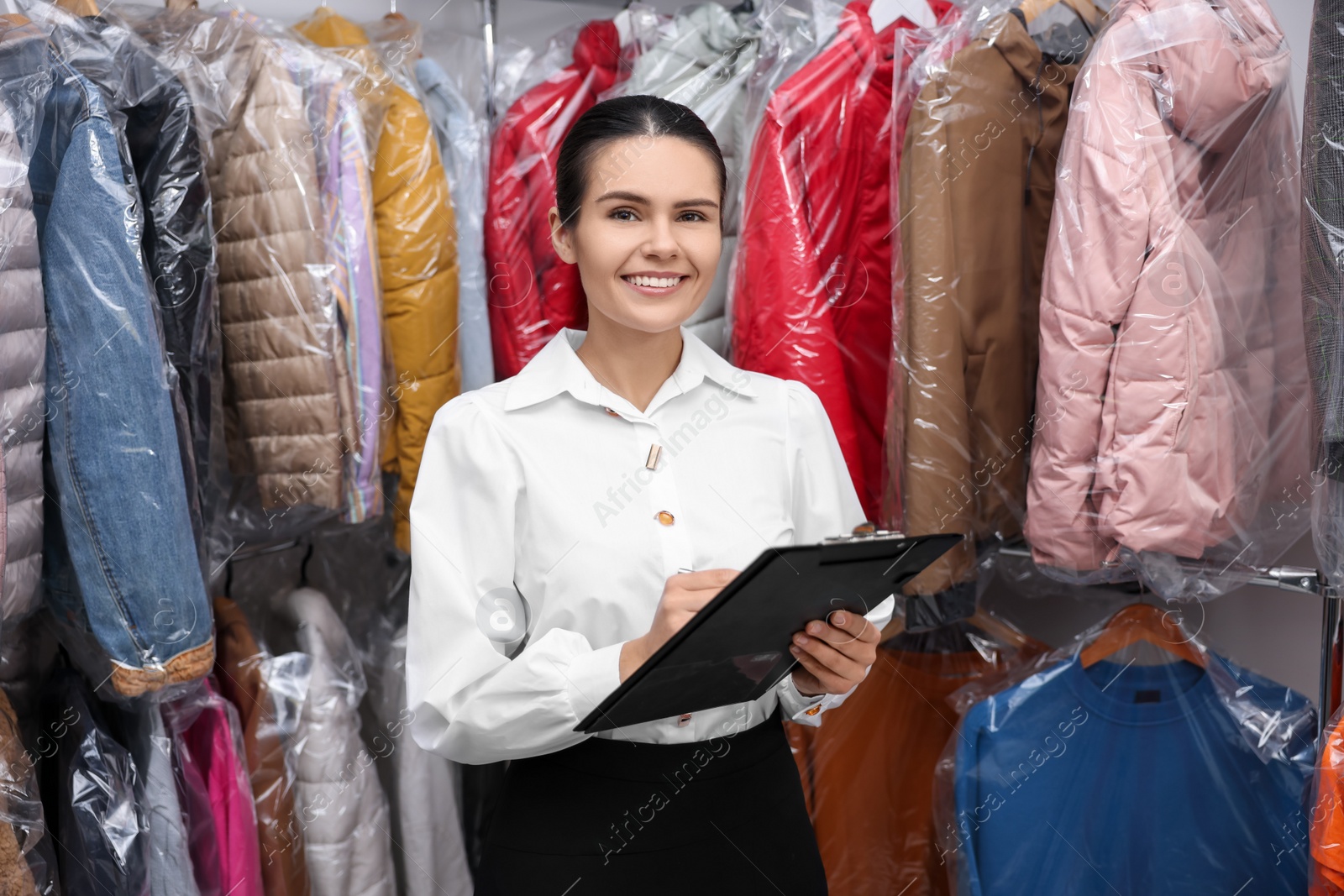 Photo of Dry-cleaning service. Happy worker holding clipboard near rack with clothes indoors