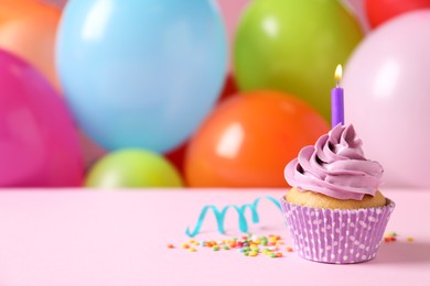 Birthday cupcake with burning candle and sprinkles on pink table against color balloons. Space for text