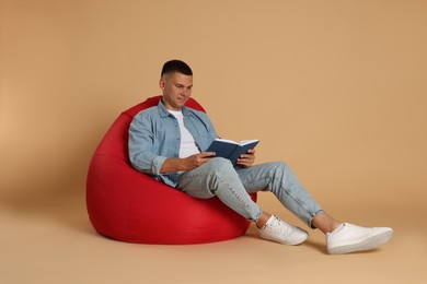 Photo of Handsome man reading book on red bean bag chair against beige background