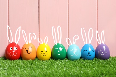 Colorful eggs as Easter bunnies on green grass against pink wooden background
