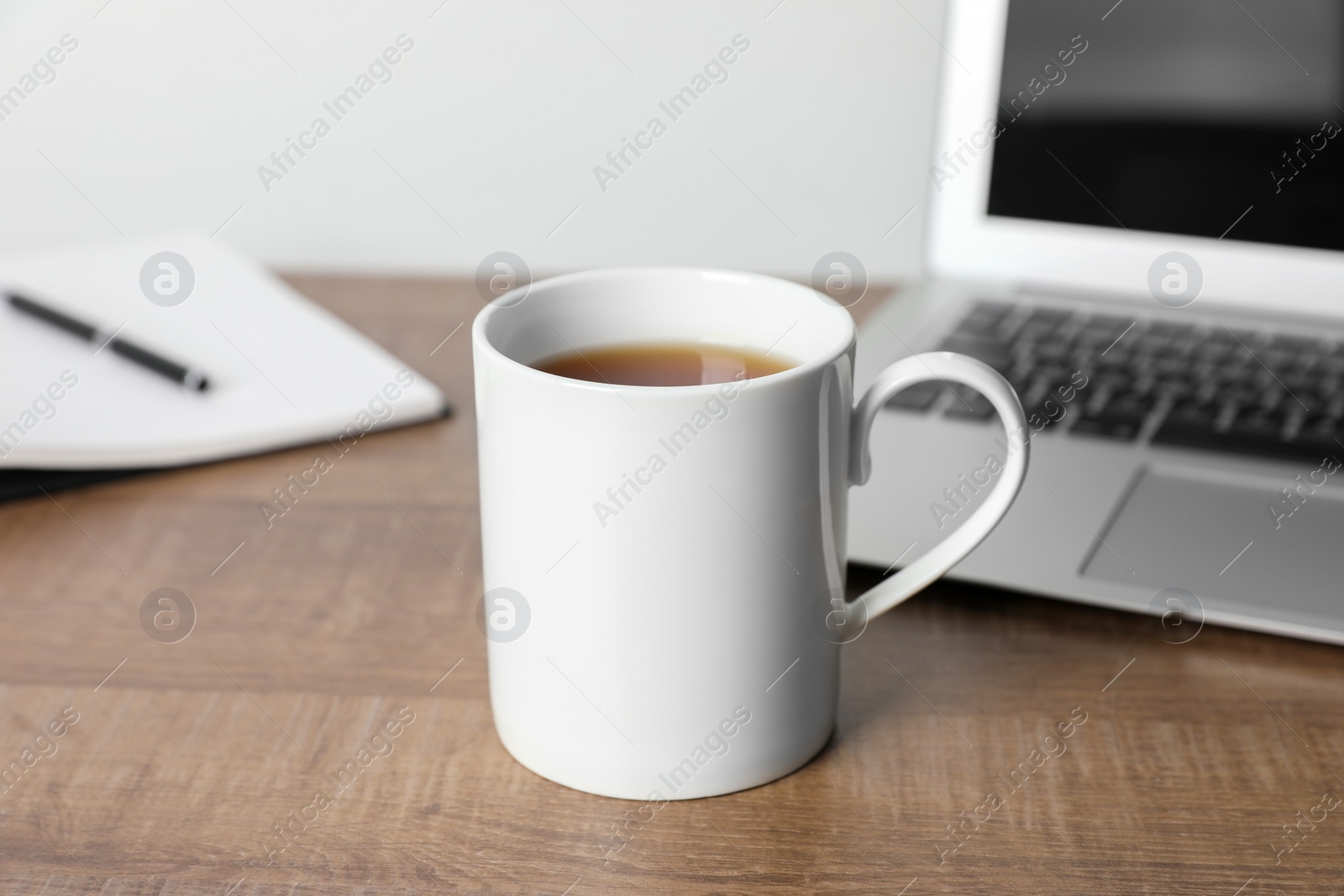 White ceramic mug with drink, notebook and laptop on wooden table at workspace Photo of White ceramic mug with drink, notebook and laptop on wooden table at workspace