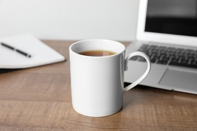 Photo of White ceramic mug with drink, notebook and laptop on wooden table at workspace