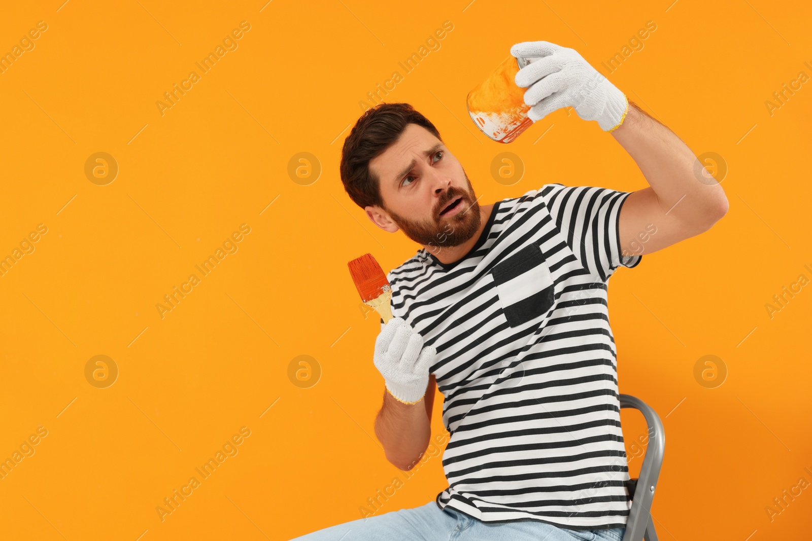 Photo of Emotional designer with painting equipment sitting on folding ladder near freshly painted orange wall, space for text