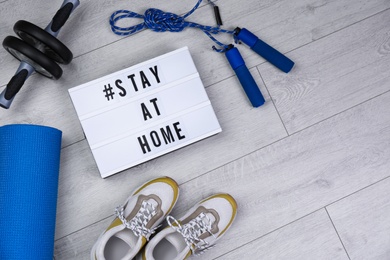 Photo of Sport equipment and lightbox with hashtag STAY AT HOME on floor, flat lay. Message to promote self-isolation during COVID‑19 pandemic