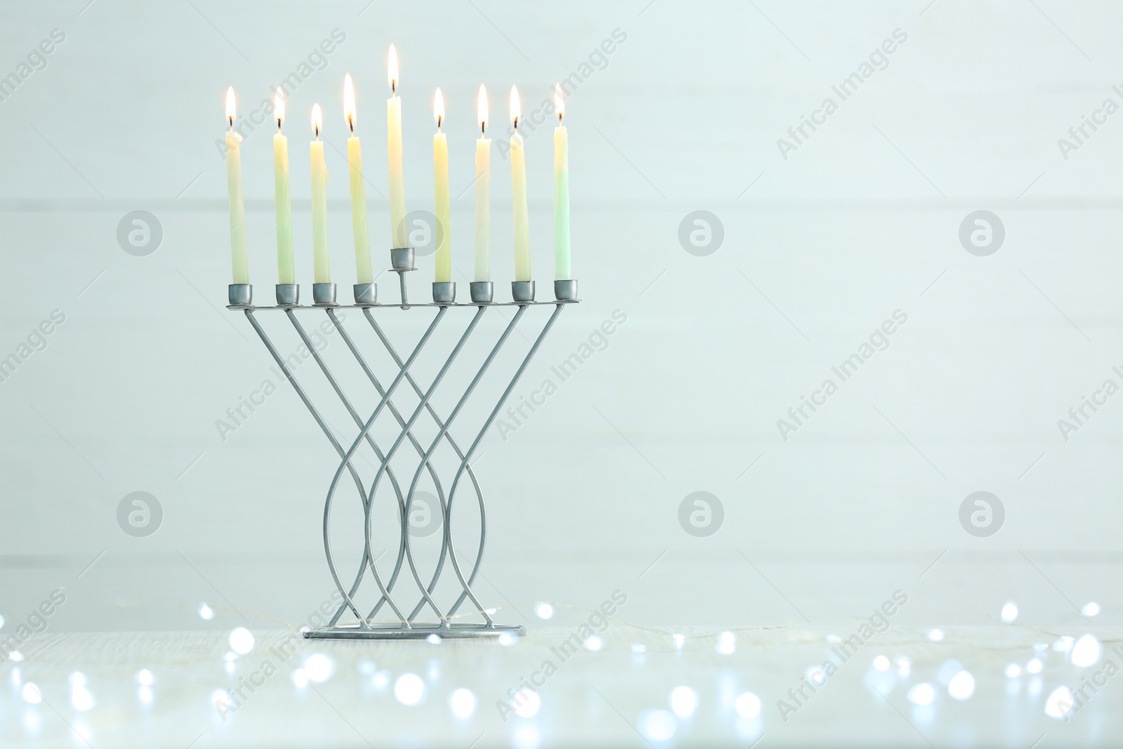 Photo of Hanukkah celebration. Menorah with burning candles and festive lights on table against light background, space for text