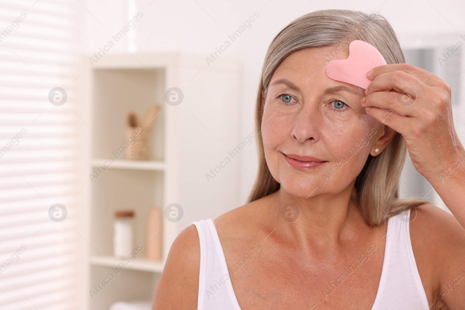 Photo of Woman massaging her face with rose quartz gua sha tool in bathroom. Space for text