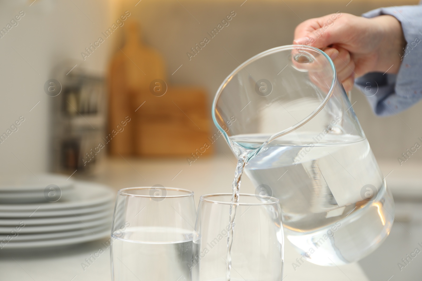 Photo of Woman pouring water from jug into glass at white table in kitchen, closeup