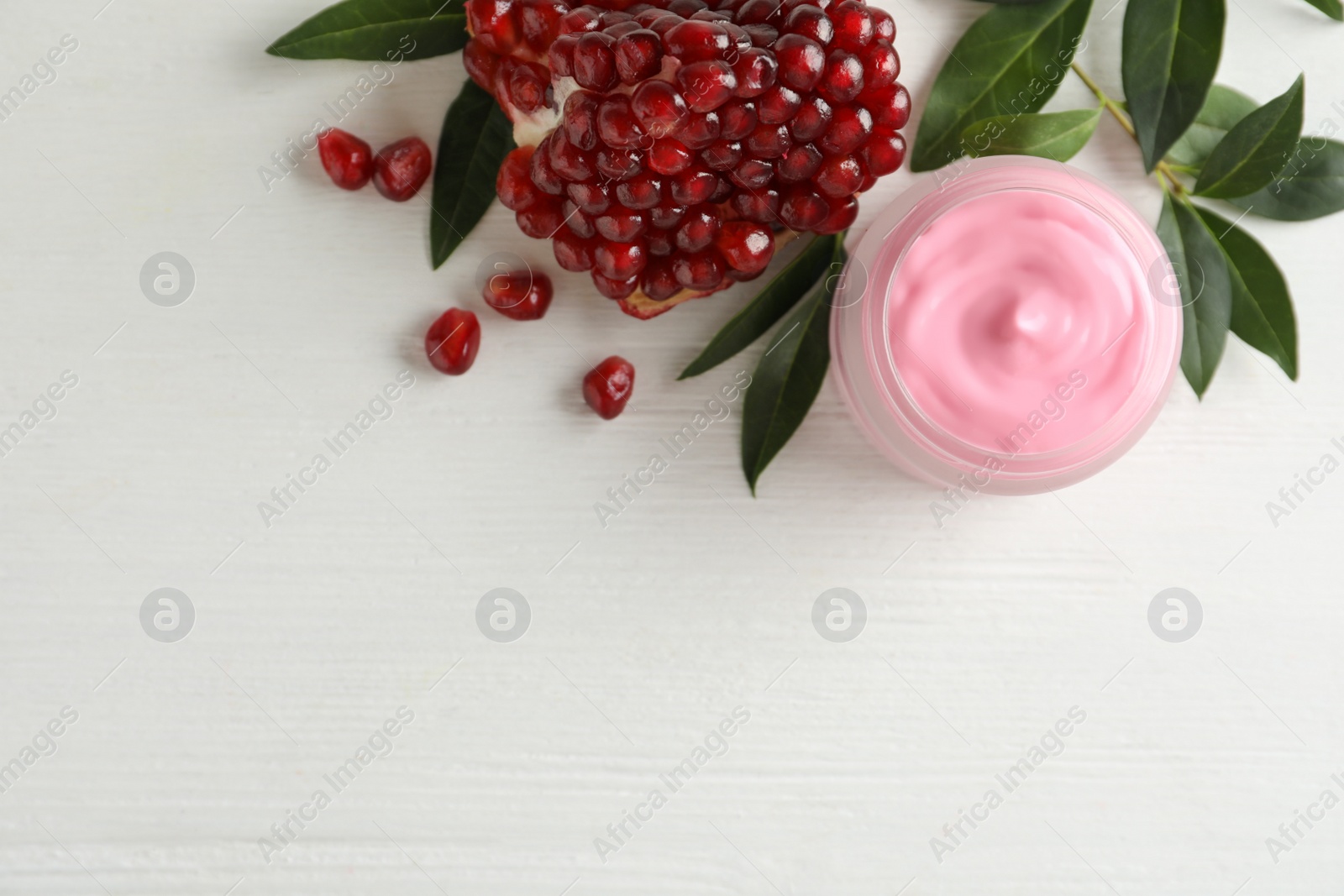 Photo of Jar with natural facial mask, pomegranate seeds and green leaves on white wooden table, flat lay. Space for text