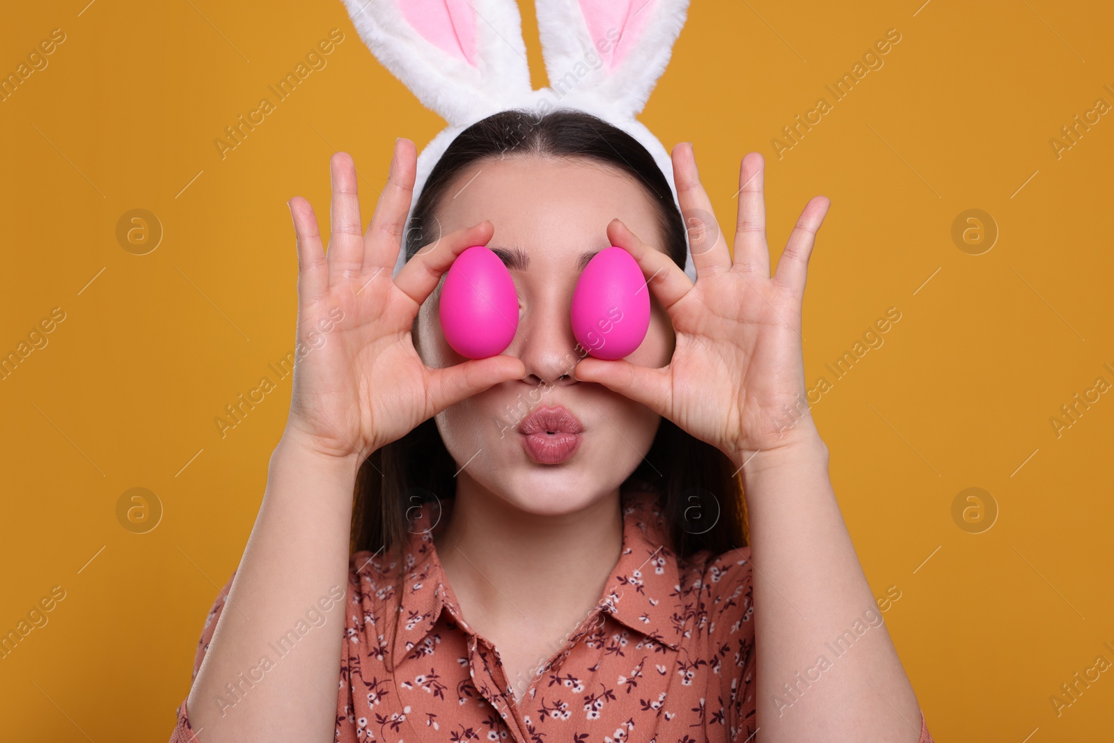 Photo of Happy woman in bunny ears headband holding painted Easter eggs near her eyes on orange background