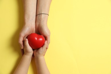 Mother and her child holding red decorative heart on pale yellow background, top view. Space for text