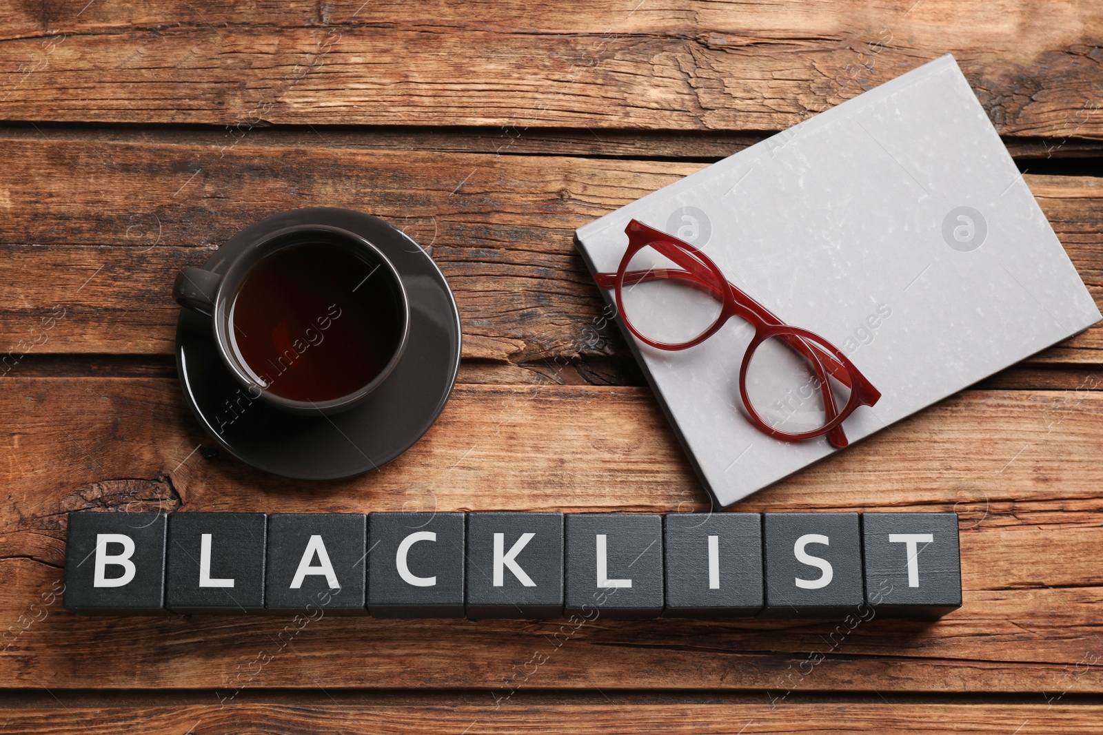 Photo of Black cubes with word Blacklist, cup of coffee and glasses on wooden desk, flat lay