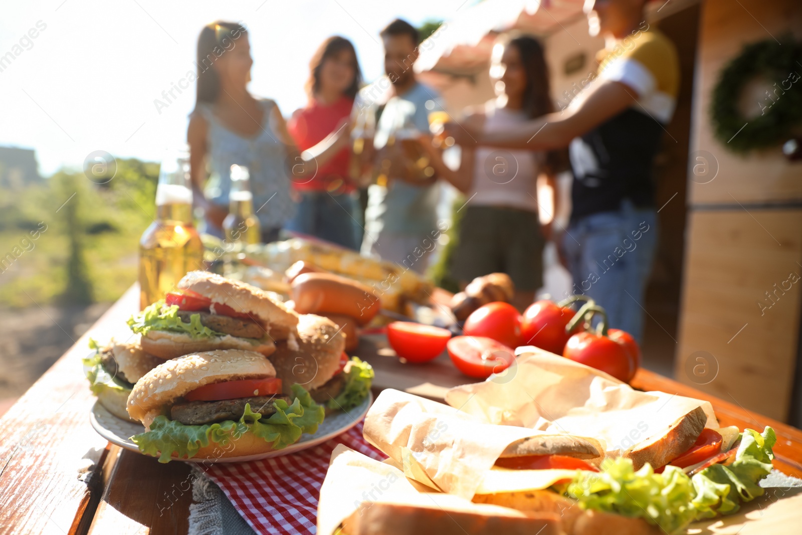 Photo of Friends toasting with bottles of drinks near motorhome, focus on table full of tasty food. Camping season