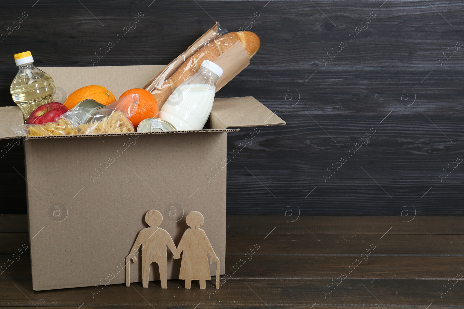 Photo of Humanitarian aid for elderly people. Cardboard box with donation food and figures of couple on wooden table, closeup
