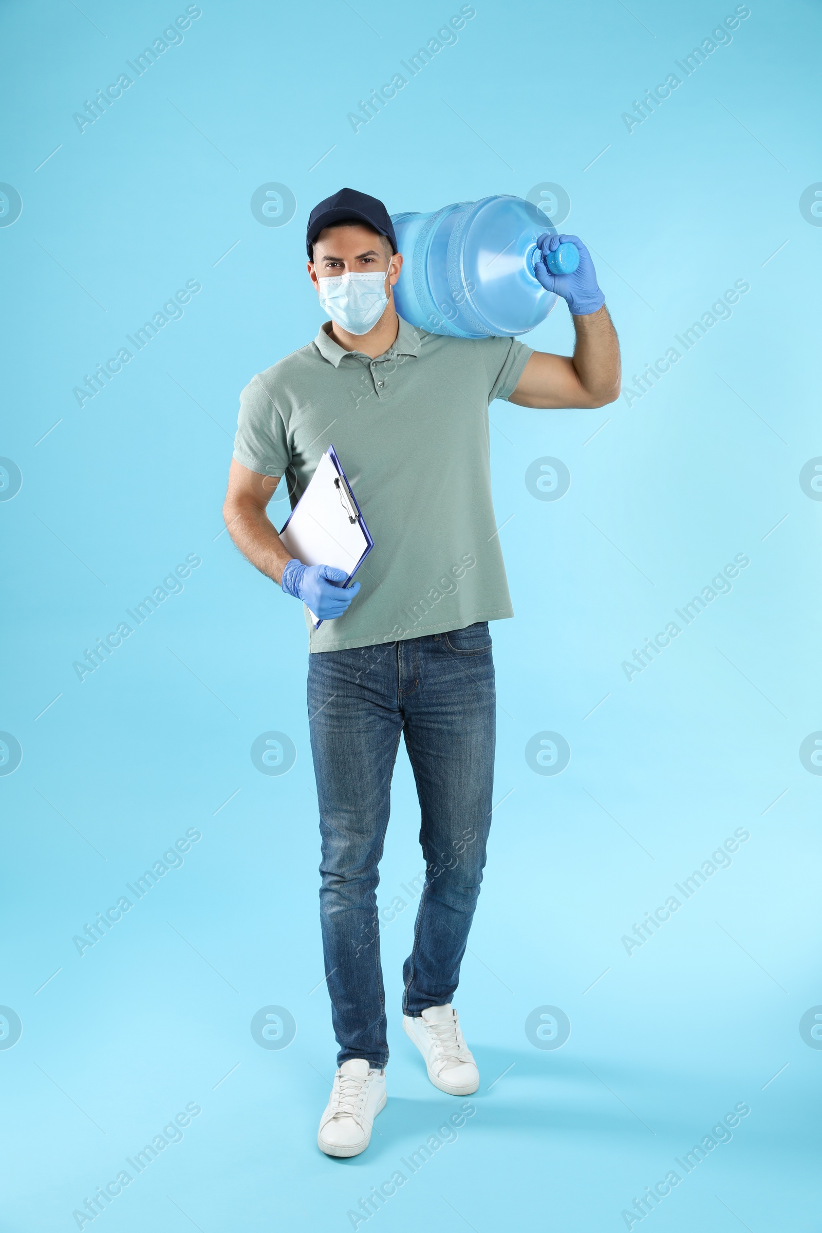 Photo of Courier in face mask with clipboard and bottle of cooler water on light blue background. Delivery during coronavirus quarantine