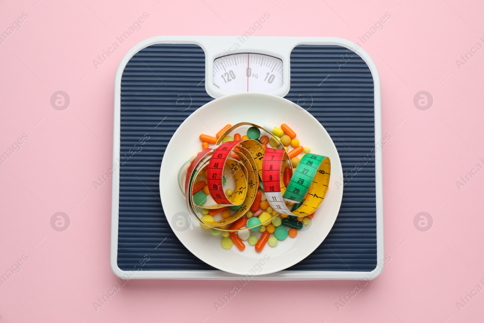 Photo of Scales with weight loss pills and measuring tape on pink background, top view