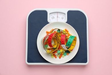 Photo of Scales with weight loss pills and measuring tape on pink background, top view