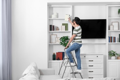 Photo of Man on metal folding ladder taking book from shelf at home. Space for text