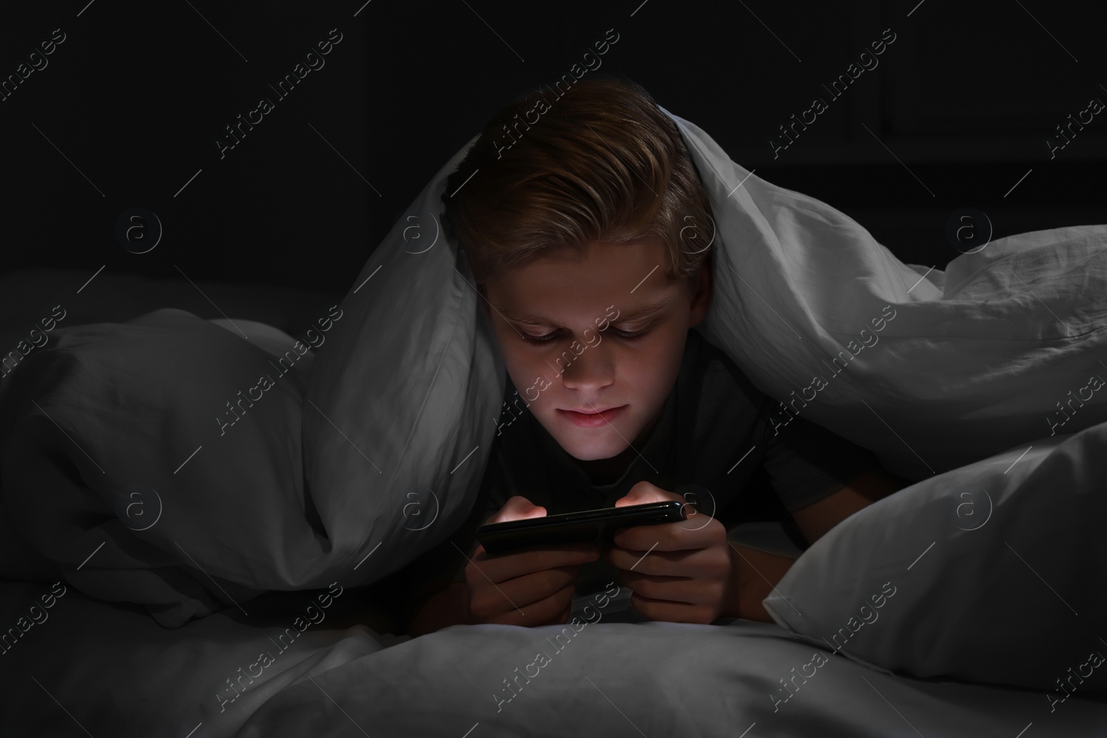 Photo of Teenage boy using smartphone under blanket on bed at night. Internet addiction