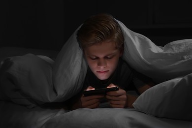 Photo of Teenage boy using smartphone under blanket on bed at night. Internet addiction