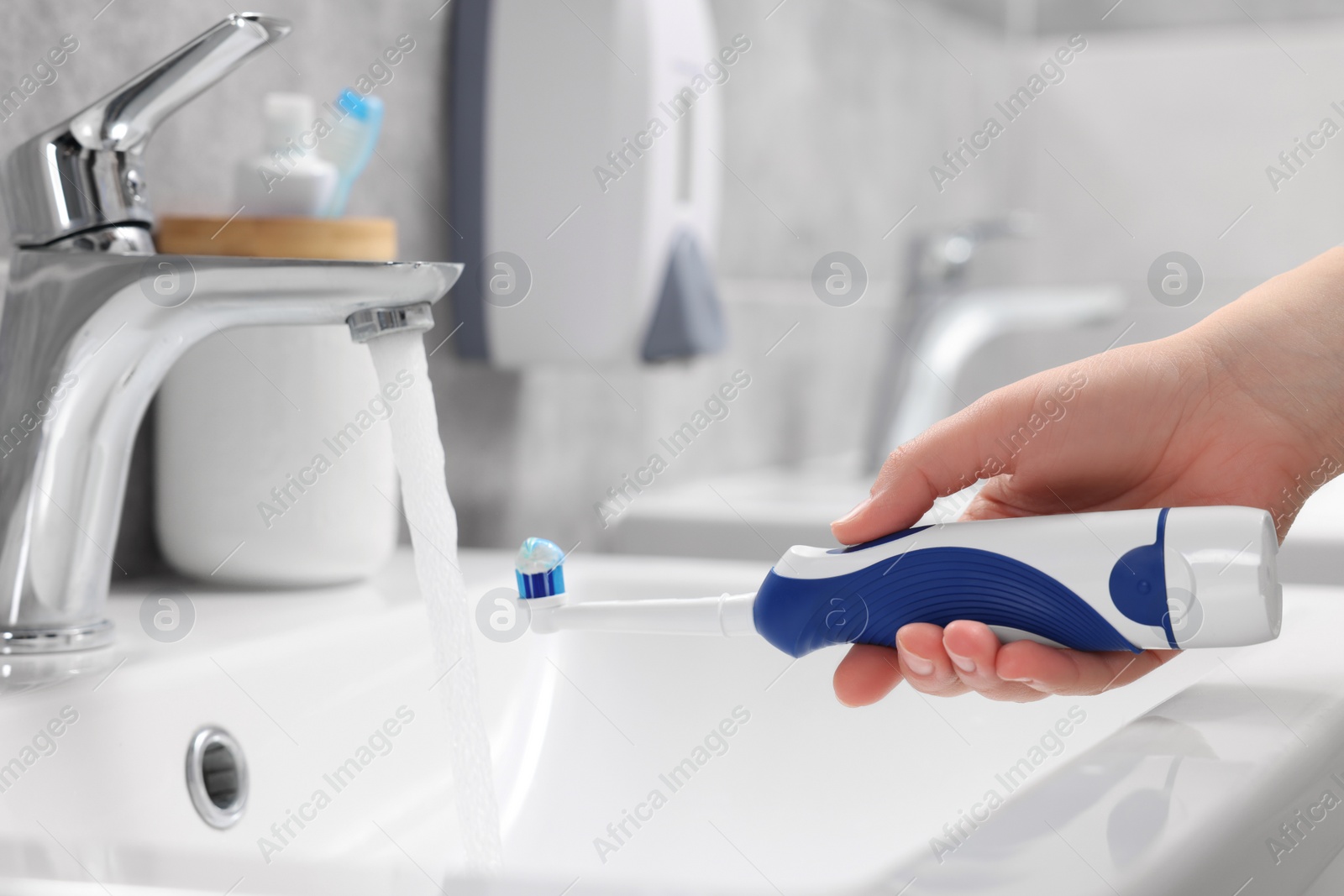 Photo of Woman holding electric toothbrush with paste near flowing water above sink in bathroom, closeup