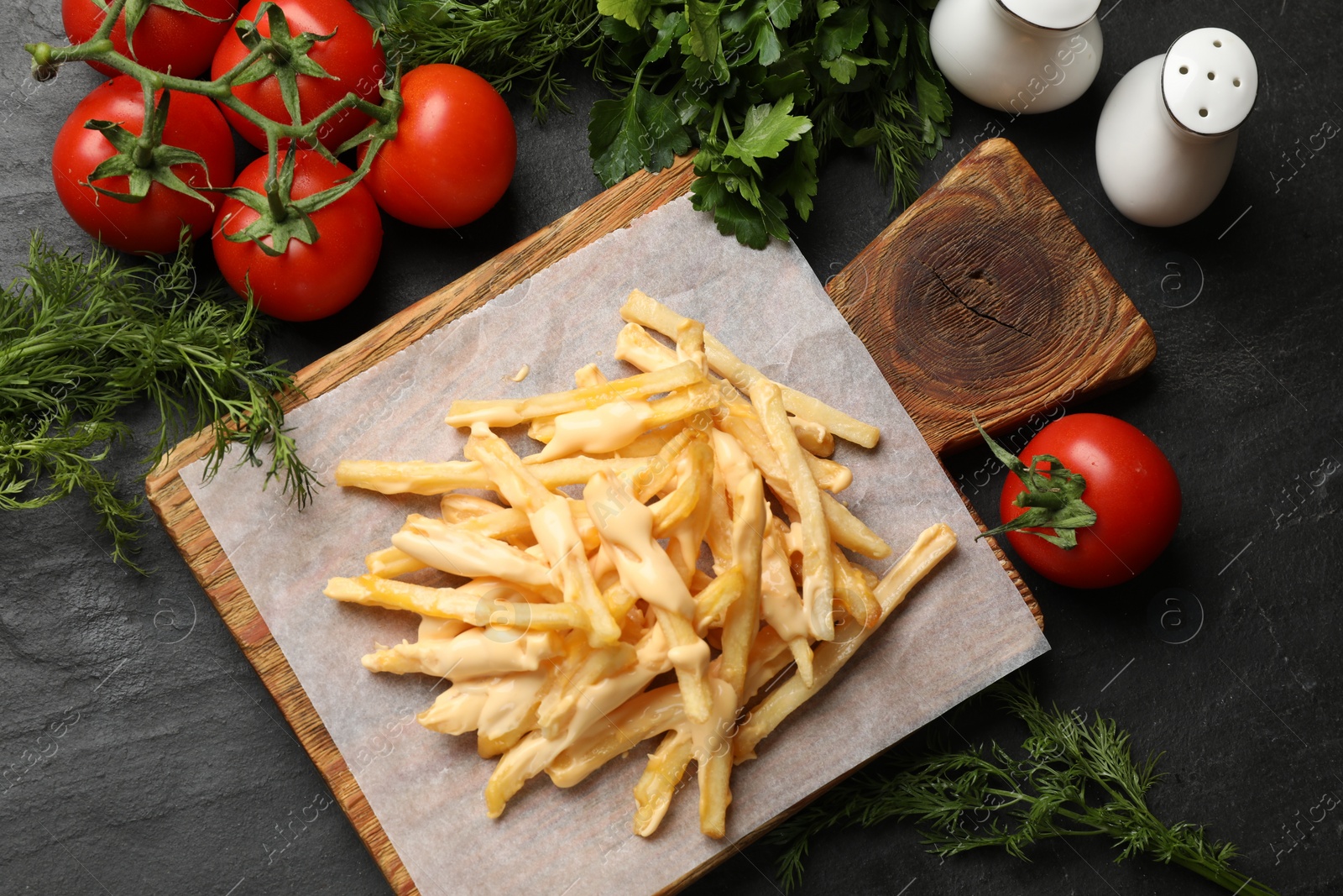 Photo of Delicious French fries with cheese sauce, tomatoes, dill and parsley on black table, flat lay