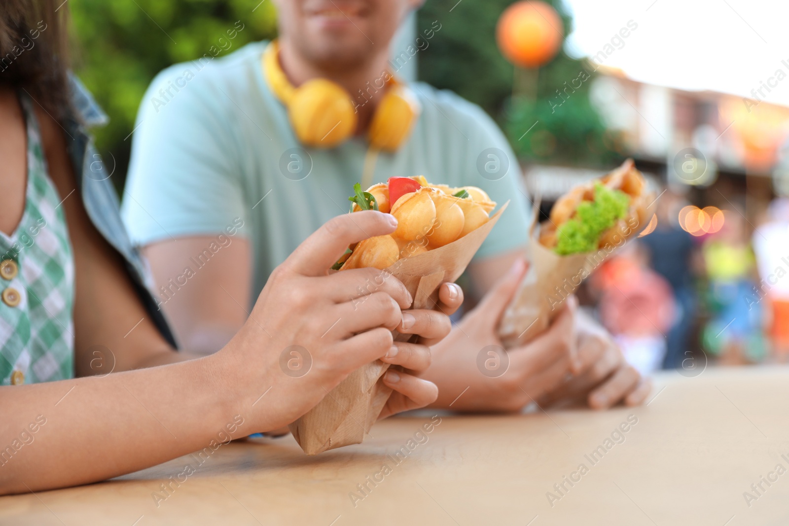 Photo of Young couple holding delicious bubble waffles with tomato and arugula at table outdoors, closeup